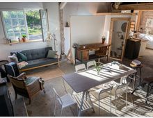 Wohnraum in einem renovierten Loft mit rustikalem Charme, bestehend aus einem grauen Sofa, Holz- und Rattansesseln, einem Esstisch mit weißen Stühlen sowie einem Schreibtisch aus Holz. Große Fenster lassen Sonnenlicht hinein, während im Hintergrund eine kleine Küche und ein gekacheltes Badezimmer sichtbar sind.