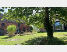 Ein Hof mit zwei roten Backsteingebäuden, eines davon hat dunkle Fensterläden und Türen, das andere besitzt eine Glasfassade. Im Vordergrund stehen ein großer Baum und Rasenflächen, umgeben von dichter Vegetation.