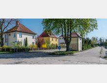 Straßenecke in einem Wohngebiet mit freistehenden Einfamilienhäusern aus hellem Putz und roten Ziegeldächern. Im Vordergrund stehen ein Baum, ein kleines Gebäude mit Graffiti und ein Verkehrsschild; im Hintergrund verläuft eine ruhige Straße.