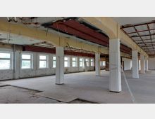 Ein leerer, sanierungsbedürftiger Innenraum mit mehreren weißen Säulen, einem beschädigten Boden und freiliegender Stahlkonstruktion in der Decke. An den Wänden befinden sich mehrere Fenster, die viel Tageslicht einlassen.