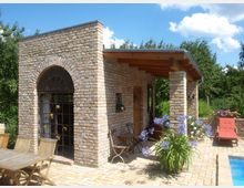 Kleines Gebäude aus Backstein mit halbrundem Fenster, einer offenen Veranda mit Holzdach und Säulen; davor stehen Holzmöbel, blühende Pflanzen und eine gepflasterte Terrasse neben einem Pool. Im Hintergrund sind grüne Bäume und ein blauer Himmel zu sehen.