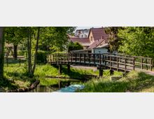 Holzbrücke über einen schmalen Bach in einer grünen Parklandschaft, umgeben von Gras und Bäumen. Im Hintergrund sind Wohnhäuser mit roten Dächern zu sehen.