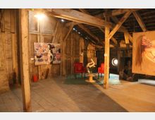 Ein rustikaler Dachboden mit Holzbalken und Holzboden, genutzt als Ausstellungsraum für Kunst. An den Wänden und auf Staffeleien sind Gemälde ausgestellt, in der Mitte stehen zwei rote Stühle und ein kleiner Tisch mit Blumen.