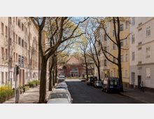 Straßenszene in einer Wohngegend mit Mehrfamilienhäusern auf beiden Seiten, Bäumen entlang des Gehwegs und geparkten Autos. Im Hintergrund steht ein Backsteinbau mit großen Fenstern und einem roten Giebel, der auf ein öffentliches Gebäude wie eine U-Bahn-Station hindeutet.