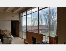 Ein unfertiger Innenraum mit großen, verglasten Fenstern, die den Blick auf eine Außenlandschaft mit Bäumen und einem roten Backsteingebäude ermöglichen. Im Raum befinden sich Kartons, Möbelteile und Baustellenmaterialien.