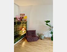 Ecke eines Wohnzimmers mit einem Ofen aus grünen, glänzenden Kacheln, darauf einige bunte Zeichnungen und eine Tischleuchte mit rosa Schirm. Vor dem Ofen stehen ein gemütlicher, gepolsterter Sessel mit rotem Muster, ein kleiner Korb in Form eines Dinosauriers und eine große grüne Zimmerpflanze.