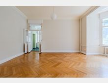 Leerer, heller Wohnraum mit Fischgrät-Parkettboden, weißen Wänden und Stuckleisten an der Decke sowie einer runden Hängelampe. Links führt eine doppelflügelige Tür in einen angrenzenden Raum; rechts sorgen große Fenster, ein Heizkörper und sichtbare Heizungsrohre entlang der Wand für Tageslicht und Details.