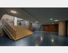 Weitläufiger Innenraum eines Gebäudes mit einer breiten Treppe aus Stein, die nach oben führt. Der Raum hat eine moderne Gestaltung mit Betonwänden, blauen Böden, runden Betonpfeilern und schlichten Deckenleuchten; Beschilderung weist auf verschiedene Bereiche hin, darunter ein Theatersaal und ein Restaurant.