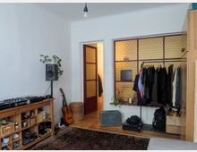 Wohnzimmer mit weiß gestrichenen Wänden, einem dunklen Teppichboden und Holzdielen. Ein offener Bereich mit gelber Glasbausteinwand trennt den Raum, im Vordergrund stehen ein Regal mit Musikequipment, eine Gitarre, sowie ein Kleiderständer mit Kleidung.