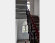 Treppenhaus mit dunkelrotem Geländer und grauen Stufen, flankiert von weißen Wänden. Zwei große Fenster mit Rundbögen lassen Tageslicht herein und bieten Blick auf eine grüne Außenanlage.