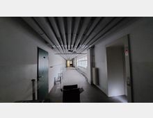 Dunkler Innenflur mit freiliegenden Rohrleitungen an der Decke, einer geschlossenen grünen Tür auf der linken Seite und einem Aufzug mit einer beleuchteten "1" rechts. Auf der rechten Wandseite befinden sich Fenster und Heizkörper, die Licht in den Raum lassen.