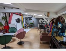 Ein modernes Fotostudio mit Holzboden und schrägen Dachfenstern. Im Vordergrund gemütliche Sessel und Tische, im Hintergrund Studioausrüstung wie Lichtständer, ein Hintergrundsystem mit verschiedenen farbigen Rollen und Accessoires auf Kleiderständern.