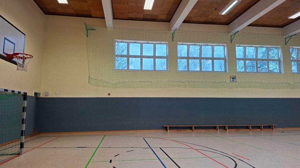 Innenansicht einer Sporthalle mit Spielfeldmarkierungen, Basketballkorb und Handballtor an der Wand. Hohe Fensterreihen mit Schutznetz, Deckenbalken mit Leuchten sowie eine lange Holzbank entlang der blaugrau gestrichenen Wand.