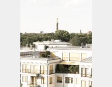 Mehrstöckiges, modernes Gebäude mit weißen Fassaden und gelben Fensterläden, umgeben von Bäumen. Im Hintergrund ist die Berliner Siegessäule mit der goldenen Statue sichtbar, vor einem dichten Waldgebiet.