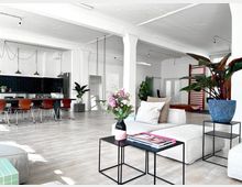 Großzügiger, offener Innenraum im Loft-Stil mit hellen Wänden und hellem Holzboden. Im Vordergrund stehen Sofas, Couchtische mit Blumen und Magazinen, im Hintergrund eine Küche mit schwarzer Wandverkleidung, Hängelampen, ein großer Esstisch und Pflanzen.