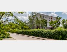 Moderne Wohngebäude mit vier Etagen, umgeben von dichter Vegetation, darunter Hecken und Bäume. Im Vordergrund verläuft ein gepflasterter Gehweg neben einer asphaltierten Straße.