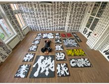 Ein Raum mit Holzfußboden und großen Fenstern, dessen Wände vollständig mit schwarzer japanischer Kalligrafie auf weißem Hintergrund bedeckt sind. Auf dem Boden liegen mehrere großformatige Kalligrafie-Gemälde in Schwarz, Weiß, Rot und Gelb, während eine Person in schwarzer Kleidung zwischen den Bildern liegt.