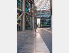 Offener Platz mit moderner Architektur, umgeben von verglasten Bürogebäuden mit roten Akzenten und sichtbaren Stahlträgern. Über dem Bereich befindet sich ein markantes, gewölbtes Dach aus Glas und Stahl, während ein einzelner Baum entlang eines gepflasterten Weges steht.