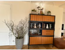 Holzregal mit schwarzem Metallrahmen in einem modern eingerichteten Raum, das mit Dekofiguren und Pflanzen geschmückt ist; daneben ein großer grauer Blumentopf mit kahlen Zweigen. Im Hintergrund helle Wände, eine weiße Tür und ein Ausschnitt einer Küche erkennbar.