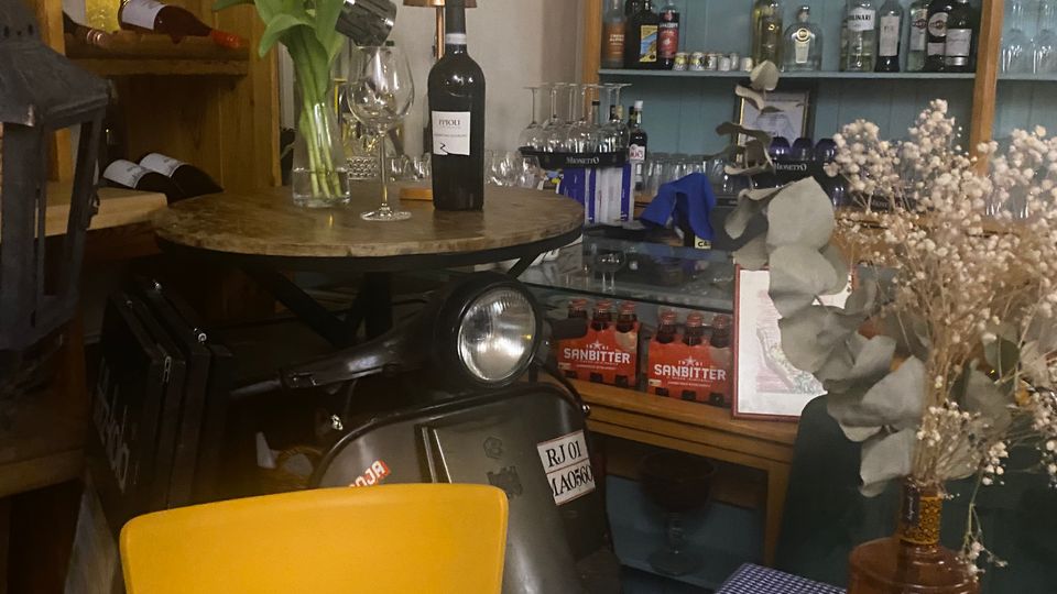 Gemütliche Bar-Ecke in einem Café mit Regalwand voller Spirituosen und Gläser sowie einem Vitrinentresen mit Getränkekisten. Im Vordergrund stehen ein Tisch mit blau-weiß karierter Tischdecke, gelbem Stuhl, Vase mit Blumen und Weinflasche; daneben ist ein Motorroller als Dekoration abgestellt.