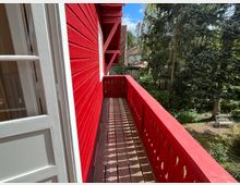 Roter Holzbalkon eines Gebäudes mit dekorativem Geländer, das in einen grünen Garten mit Bäumen und Sträuchern blickt; im Vordergrund ein weiß gerahmtes Fenster.