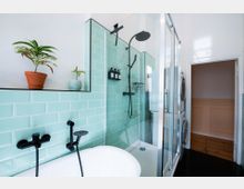Modernes Badezimmer mit einer Badewanne und einer begehbaren Dusche, die mit mintfarbenen Fliesen gestaltet ist. Schwarze Armaturen, zwei kleine Zimmerpflanzen auf einer Ablage sowie eine Waschmaschine und ein Trockner sind ebenfalls sichtbar.