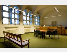 Innenraum eines Gerichtssaals mit hohen Fenstern, durch die Tageslicht einfällt. Der Raum verfügt über eine gewölbte Decke, einen Tisch mit Stühlen in der Mitte, eine hölzerne Richterbank im Hintergrund sowie eine Holzbank im Vordergrund.