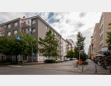 Straßenszene in einer Wohngegend mit mehrstöckigen Wohngebäuden, Bäumen entlang der Straße und parkenden Autos. Am Straßenrand befinden sich ein Zebrastreifen, ein Kiosk und Straßenbeschilderungen mit gepflastertem Gehweg.