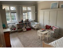 Helles Kinderzimmer mit großen Fenstern, die von weißen Fensterläden eingerahmt sind. Der Raum enthält ein Gitterbett, ein Bücherregal voller Bücher und Spielzeug, ein kleines Sofa mit gemustertem Bezug, einen beigefarbenen Teppich sowie einen kleinen Tisch mit zwei Stühlen.