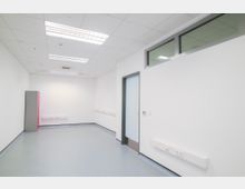 Ein leerer, rechteckiger Raum mit weißen Wänden, grauem Boden und einer abgehängten Decke mit eingebauten Leuchtstoffröhren. An der rechten Wand befindet sich eine Tür mit Glasfenster sowie Steckdosenleisten, und in der linken Ecke steht ein schmaler, grauer Schrank mit einer roten Seite.