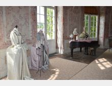 Eleganter Raum mit barockem Dekor, ausgestellten historischen Kostümen auf Schaufensterpuppen und einem schwarzen Flügel, auf dem kleine Figuren und Dekorationsobjekte platziert sind; große Fenster lassen Tageslicht herein.