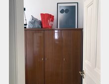 Holzschrank mit glänzender Oberfläche, darauf stehen eine schwarz-weiß karierte Tasche, eine rote Tragetasche mit Aufschrift und ein gerahmtes Poster an der weiß gestrichenen Wand. Ein teilweise geöffneter Türrahmen mit goldfarbenem Türgriff ist rechts sichtbar.