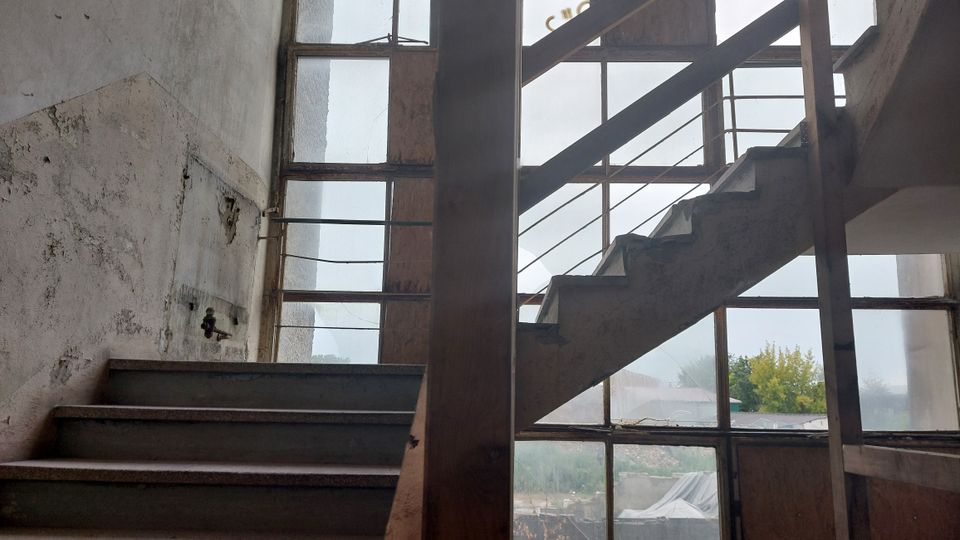 Treppe in einem verlassenen Gebäude mit verfallenen Wänden und großen Fensterfronten aus Glas, die den Blick auf eine bewachsene Außenlandschaft freigeben. Die Metallgeländer und die sichtbaren Abnutzungsspuren deuten auf einen vernachlässigten Zustand hin.