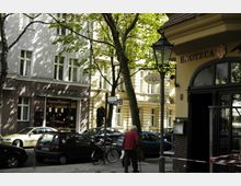 Straßenszene in einer städtischen Umgebung mit Bäumen, einem grauen Gebäude mit einer Bäckerei im Erdgeschoss und einem gelben Gebäude mit einem "Enoteca"-Schild. Autos parken entlang der Straße, und eine Person mit roter Jacke geht zu Fuß mit einem Gehstock vorbei.