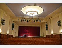 Auditorium mit einer Bühne, auf der ein schwarzer Flügel und ein Mikrofon stehen; der Hintergrund besteht aus einem roten Vorhang. Der Raum hat beige Wände, eine moderne Deckenleuchte, Reihen von Sitzplätzen und technische Ausstattung wie Lautsprecher und Bühnenbeleuchtung.