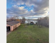 Ein weitläufiger Garten mit kurzgemähter Wiese und vereinzelten Bäumen führt zu einem kleinen See. Links steht ein flach gebautes, rosafarbenes Haus mit Garageneinfahrt, während im Hintergrund Wolken den Himmel bedecken.