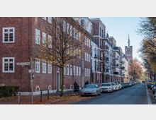 Straßenszene in einem Wohngebiet mit Backsteinfassaden und modernen Gebäuden mit Balkonen. Im Hintergrund ist der Turm einer Kirche zu sehen, während die Straße von Bäumen gesäumt und mit parkenden Autos belegt ist.