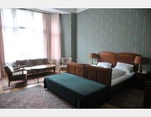 Ein klassisch eingerichtetes Schlafzimmer mit einem Doppelbett aus Holz, flankiert von Nachttischen mit Lampen. Im Raum befinden sich außerdem eine Sitzecke mit Sofa, Sesseln und einem ovalen Couchtisch sowie große Fenster mit Vorhängen, ein grünes Tapetenmuster und Teppiche auf Holzboden.