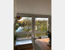 Ein Raum mit großen Fenstern, die den Blick auf eine herbstliche Landschaft mit Bäumen und einem See freigeben. Im Vordergrund stehen ein Holztisch mit Blumenvasen und Fotos sowie ein Heizkörper unter der Fensterbank.