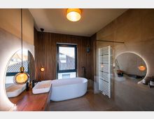 Modernes Badezimmer mit einer freistehenden weißen Badewanne, einem großen, beleuchteten runden Spiegel und Holzvertäfelung an der Wand. Es gibt ein großes Fenster, eine weiße Handtuchheizung und eine hängende Designerleuchte über einem Waschtisch mit rechteckigem Waschbecken.