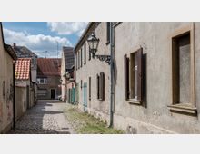Schmale Kopfsteinpflastergasse mit älteren, verputzten Häusern auf beiden Seiten, teilweise mit Holztüren und Fensterläden. Laternen im traditionellen Stil hängen an den Gebäudewänden, im Hintergrund sind Häuser mit roten Ziegeldächern zu sehen.