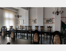 Ein Versammlungsraum mit langen, dunklen Holztischen und gepolsterten, verzierten Stühlen, eingerahmt von weißen Wänden. An den Wänden hängen Bilderrahmen, und zwei dekorative Kronleuchter hängen von der Decke.