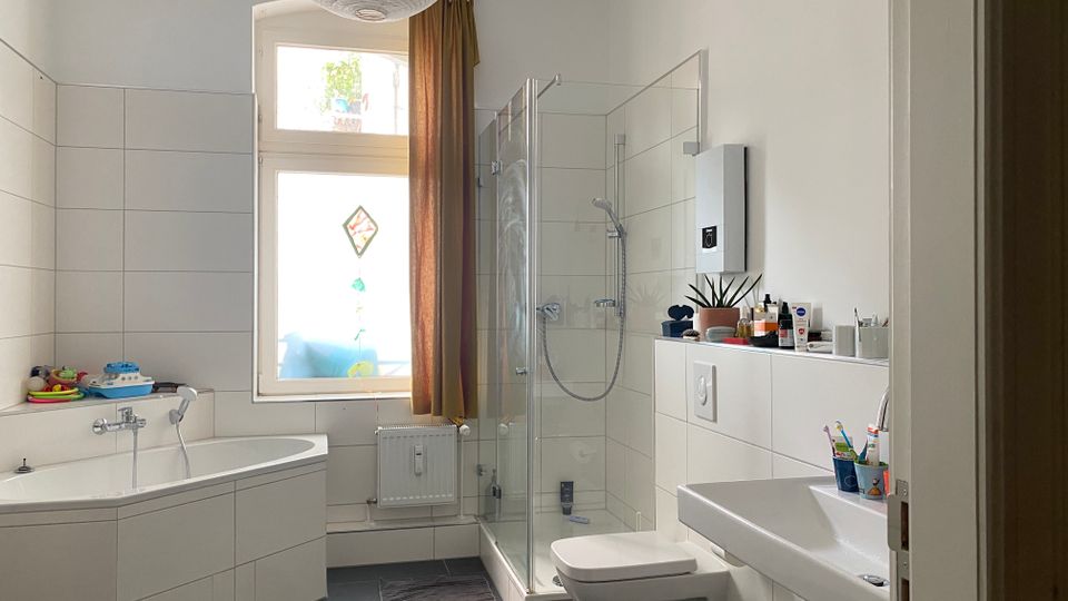 Helles Badezimmer mit weißen Wandfliesen und dunklem Boden, ausgestattet mit Eckbadewanne, verglaster Dusche, wandhängendem WC und großem Waschbecken. Ein großes Fenster mit Vorhang bringt Tageslicht hinein; auf einer Ablage stehen Pflegeprodukte und eine Pflanze.