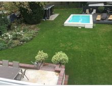 Blick auf einen gepflegten Garten mit einer großen Rasenfläche, einem rechteckigen Pool am oberen Rand, einer angrenzenden Terrasse mit Liegestühlen sowie einem bepflanzten Blumenbeet und einem kleinen Gartenschuppen. Im Vordergrund ist eine Steinterrasse mit einem Tisch, Stühlen und einer Gießkanne zu sehen.