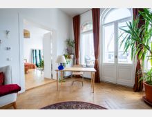 Heller Wohnraum mit einem Schreibtisch aus Holz, einer Lampe und Pflanzen, vor großen weißen Doppelfenstern mit bogenförmigen Oberlichtern und braunen Vorhängen. Im Hintergrund ein angrenzendes Zimmer mit Holzboden und einem Bett.