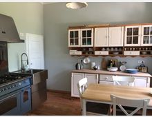 Helle Landhausküche mit einem Holzfußboden, einem großen Gasherd mit Edelstahlspüle, cremefarbenen Schränken mit Glasfronten und Regalen sowie einem Holztisch mit weißen Stühlen im Vordergrund. Verschiedene Küchenutensilien, Geschirr und Gewürzflaschen sind auf der Arbeitsfläche und in den Regalen angeordnet.