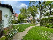Vorgarten einer Wohnsiedlung mit Rasenfläche, Blumenbeeten und einem gepflasterten Weg, der zu einem Metalltor führt. Im Hintergrund stehen Bäume, Hecken, Häuser mit klassischen Dachformen sowie ein Haus mit Solarpanels.
