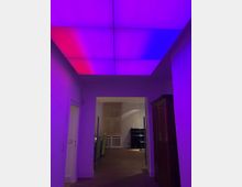 Ein Innenraum mit farbig beleuchteter Decke in Lila- und Rottönen, der in einen angrenzenden Raum mit Holzboden und Möbeln führt. Im Vordergrund steht ein Schrank, während im hinteren Bereich ein heller, schlicht möblierter Raum sichtbar ist.