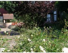 Ein idyllischer Garten mit einem alten Brunnen aus Ziegelsteinen, umgeben von blühenden Sträuchern und Blumen. Im Hintergrund sind eine gepflasterte Fläche, ein Zierbaum und ein Gebäude mit weißen Türen und roten Dächern zu sehen.
