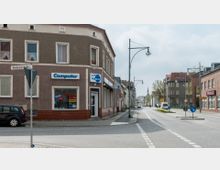 Straßenszene mit einer Kreuzung, an der sich ein zweigeschossiges Eckgebäude mit brauner Fassade und Geschäften im Erdgeschoss befindet, darunter ein Computerladen. Rechts verläuft eine Hauptstraße mit Wohn- und Geschäftsgebäuden, am Horizont ist ein Kirchturm sichtbar.